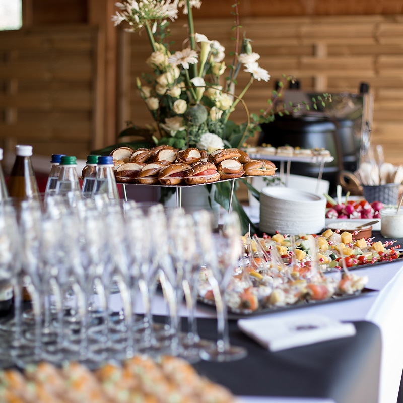 Table de réception : mini-sandwiches, brochettes colorées, mignardises et bouteilles. Verres à pied alignés, grande composition florale blanche.