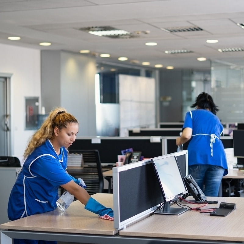 Employées de nettoyage en uniforme bleu désinfectent un bureau moderne. L'une essuie un poste de travail, l'autre nettoie à l'arrière-plan.